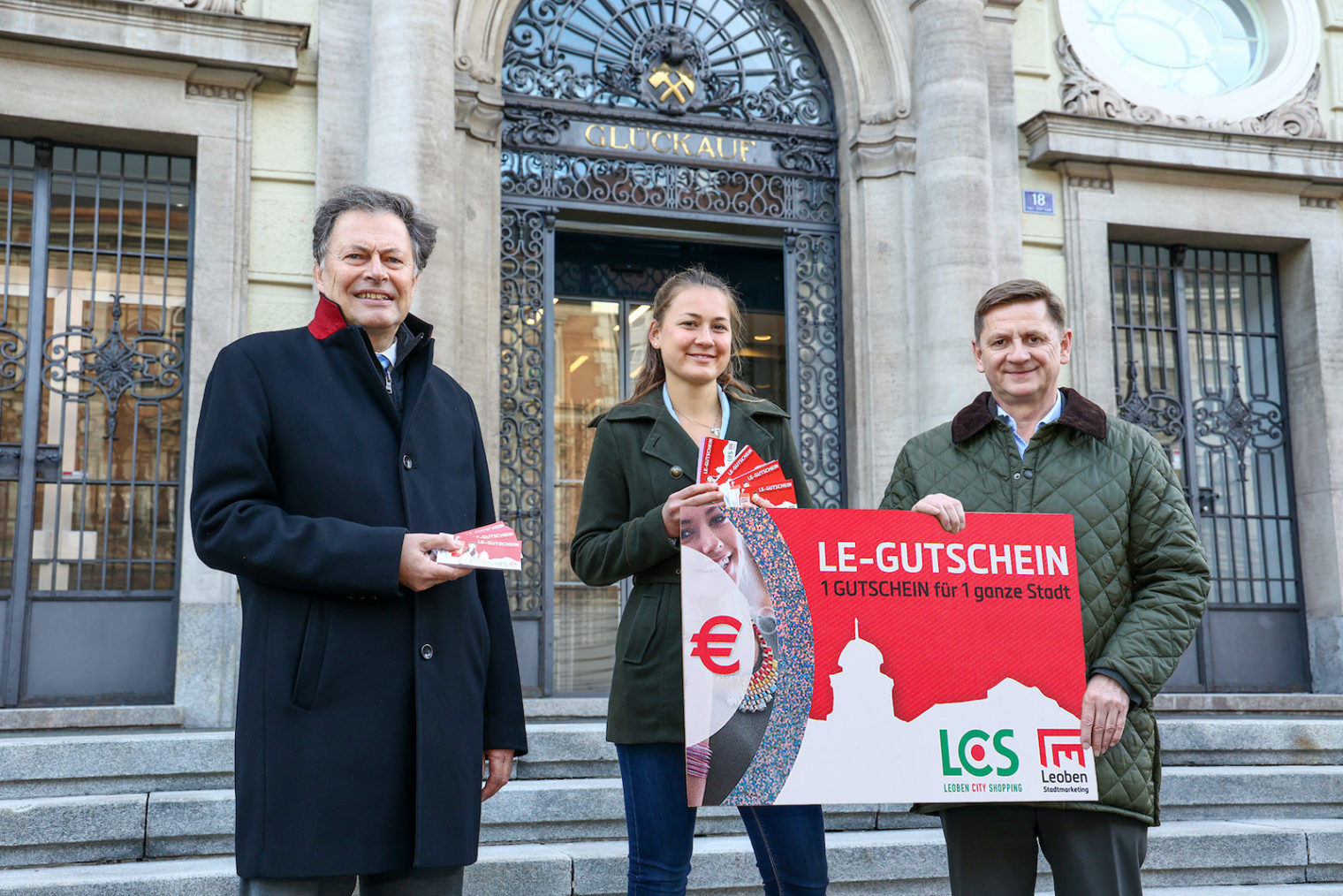 Vor dem Eingang der Montantuniversität Leoben stehen Magnifizenz Wilfried Eichlseder (Rektor der Montanuniversität Leoben) mit Leoben-Gutscheinen, Julia Brandstetter (Vorsitzende der Hochschülerschaft an der Montanuniversität Leoben) mit Leoben-Gutscheinen. Sie hält gemeinsam mit Bürgermeister Kurt Wallner Plakat zu den Gutscheinen mit dem Text: 