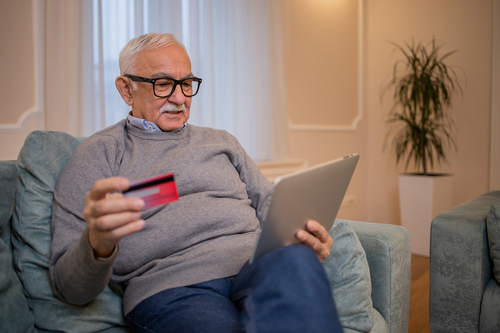 Älterer Mann sitzt mit Bankkarte in der Hand vor dem Tablet