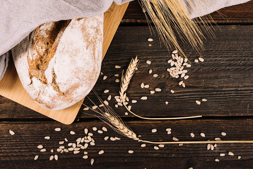 Symbolbild: Detailansicht eines Laibes Brot auf einem Holzbrett, auf dem Tisch liegen Getreidekörner und Halme