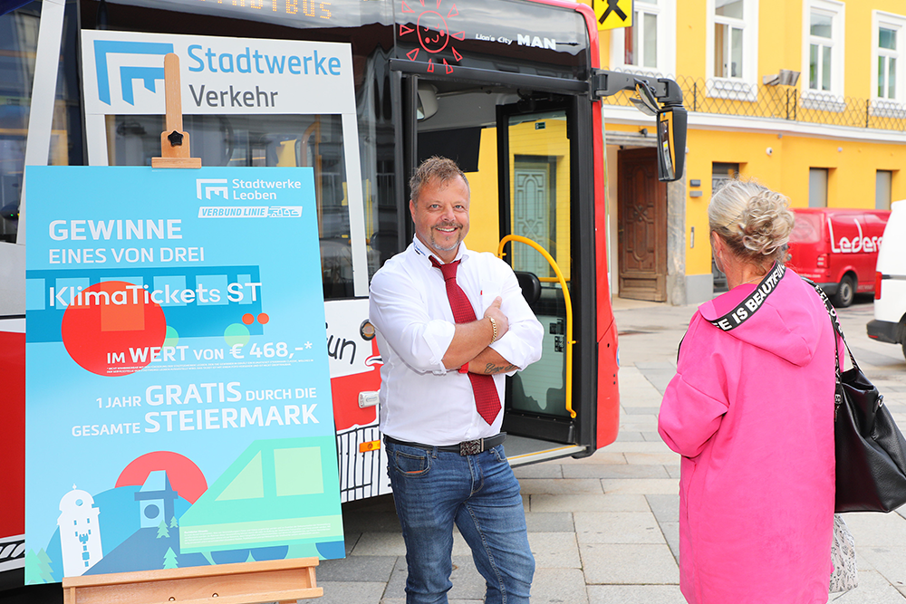 Busfahrer der Stadtwerke Leoben vor einem Bus und Infotafel zum Klimaticket-Gewinnspiel