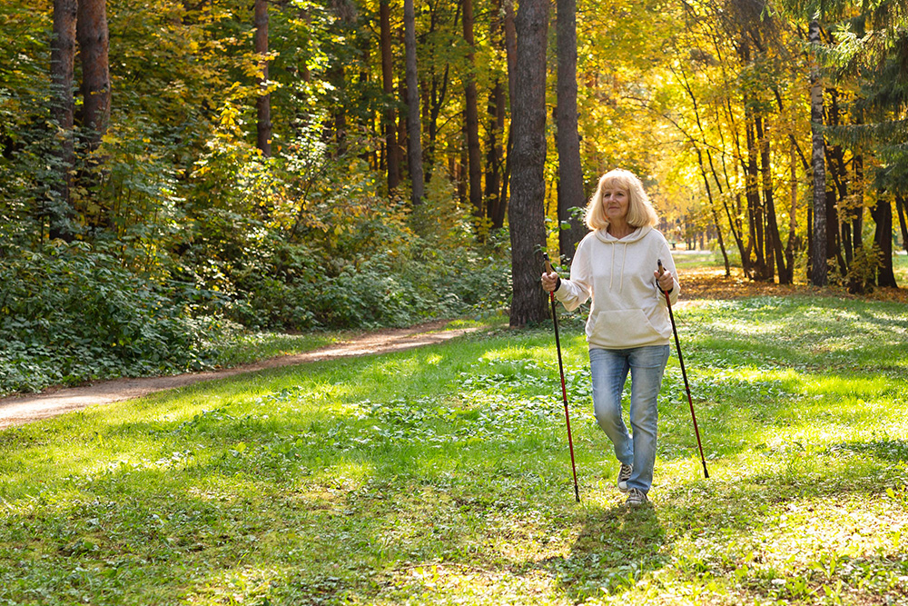 Eine Seniorin in Jeans und Pullover spaziert mit Walking-Stöcken im herbstlichen Wald