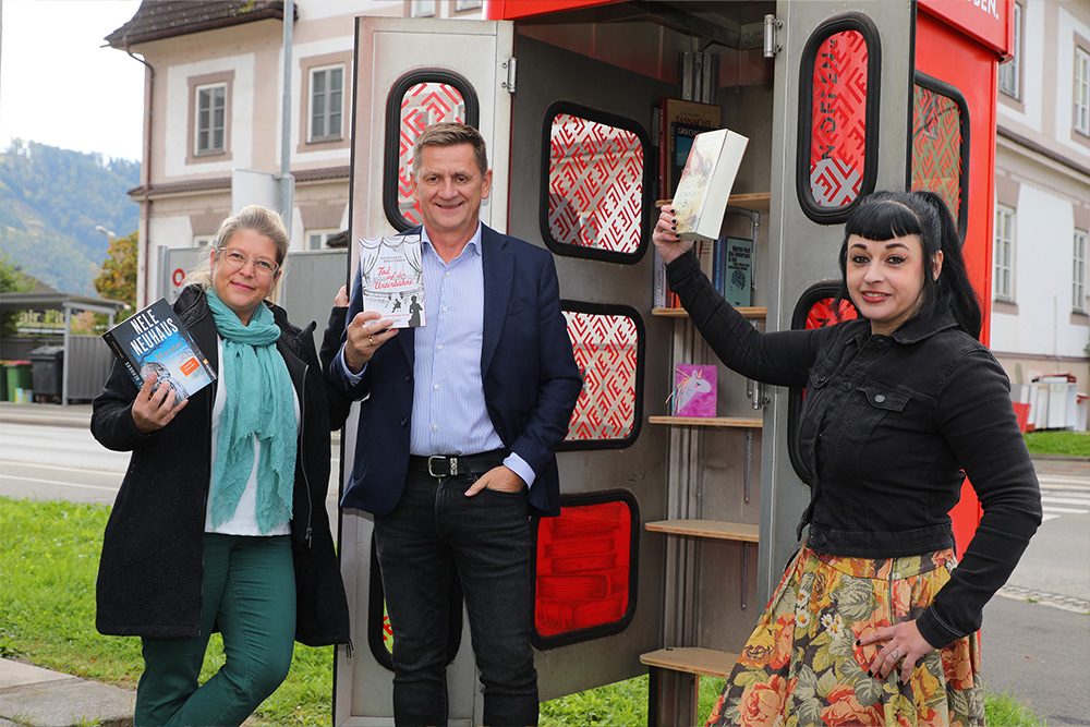Drei Personen vor einer neuen Bücherzelle, jede ein Buch in der rechten Hand hochhebend
