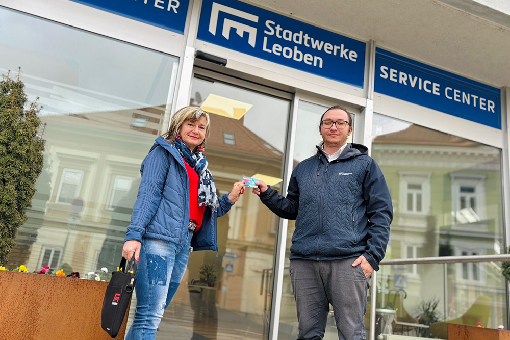Eine Dame und ein Herr stehend vor dem Stadtwerke Leoben Service Center halten gemeinsam ein Klimaticket in den Händen.