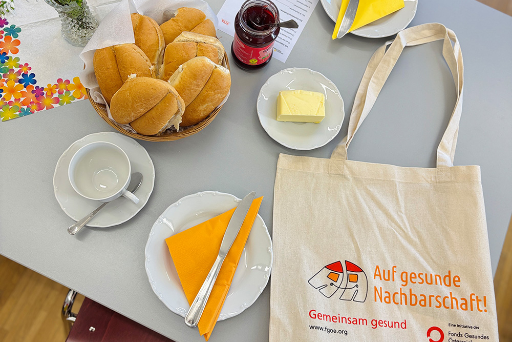 Gedeckter Frühstückstisch mit Semmeln, Butter und Marmelade und einer Stofftasche mit der Aufschrift Auf gesunde Nachbarschaft