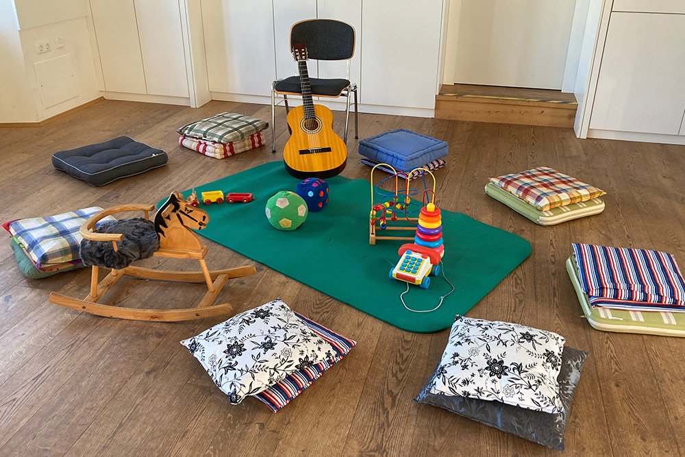 Spielsachen und Gitarre in der Mitte auf einer Matte und rundherum ein Sessel und Sitzpölster am Boden liegend