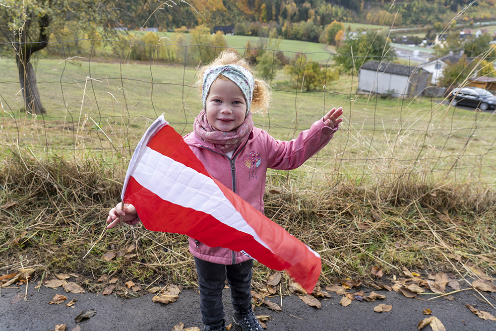 Kleines rothaariges Mädchen mit österreichischer Fahne in der Hand, steht vor einem Weidezaun
